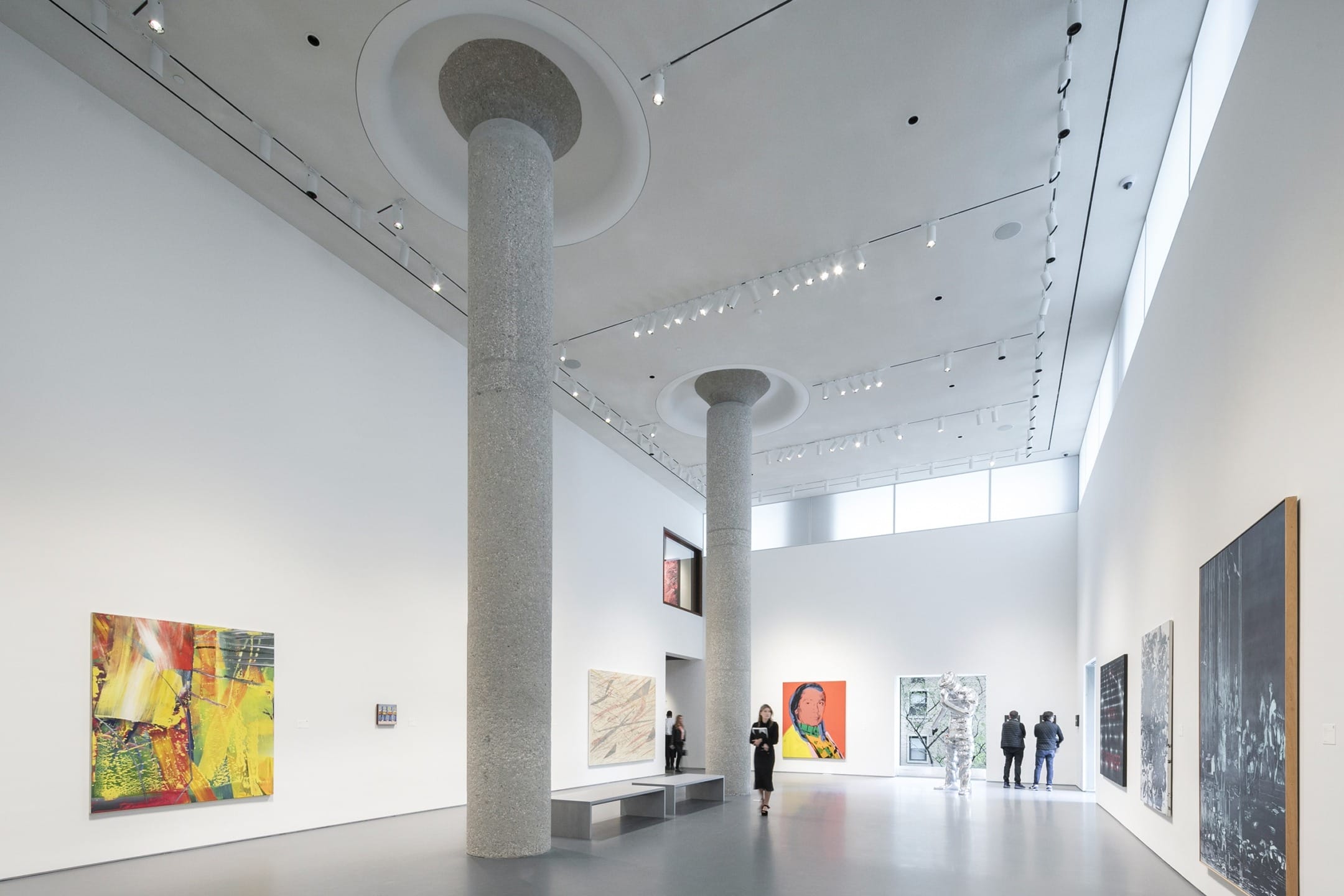 Spacious art gallery with tall columns, visitors viewing various artworks on display, modern lighting from ceiling fixtures.