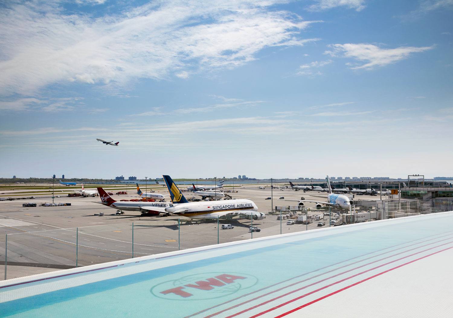 View of an airport runway with several airplanes parked and one taking off under a blue sky.