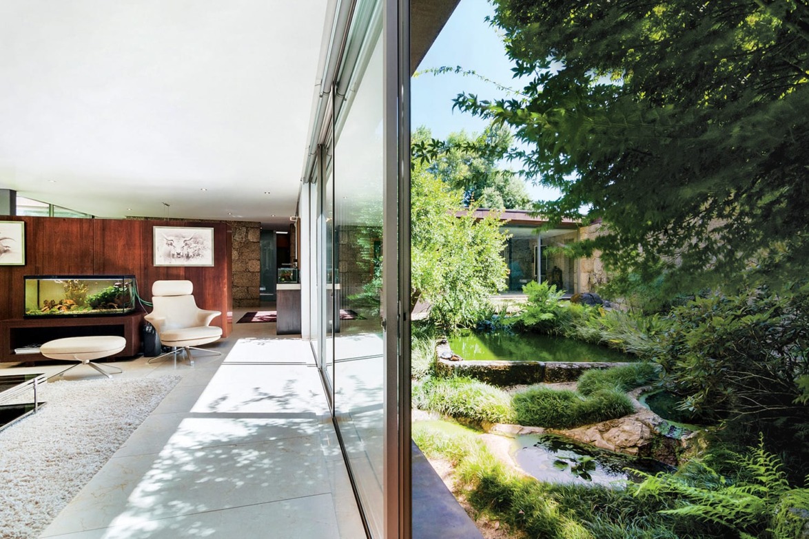 Modern living room with large windows overlooking a lush garden, featuring a white chair and fish tank inside.