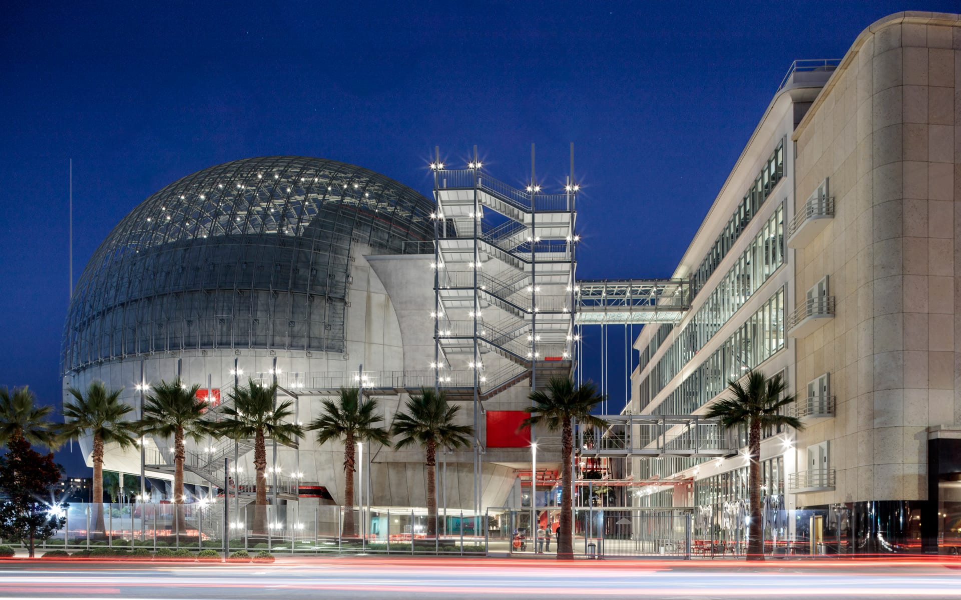 Modern building at night with a large, dome-shaped structure and palm trees lining the foreground.