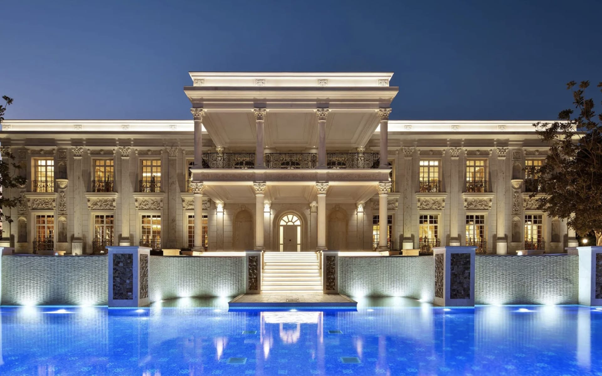 Elegant neoclassical mansion with illuminated facade, grand entrance, and a reflective pool at dusk.
