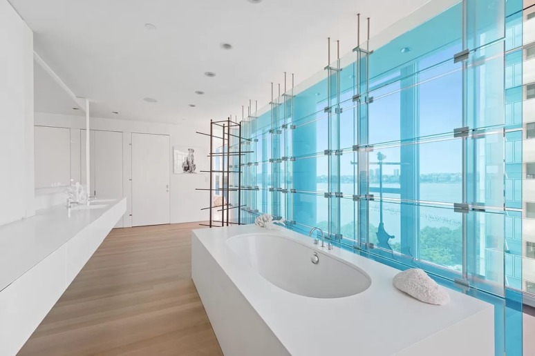 Modern bathroom with floor-to-ceiling windows, freestanding tub, and view of a body of water and cityscape.