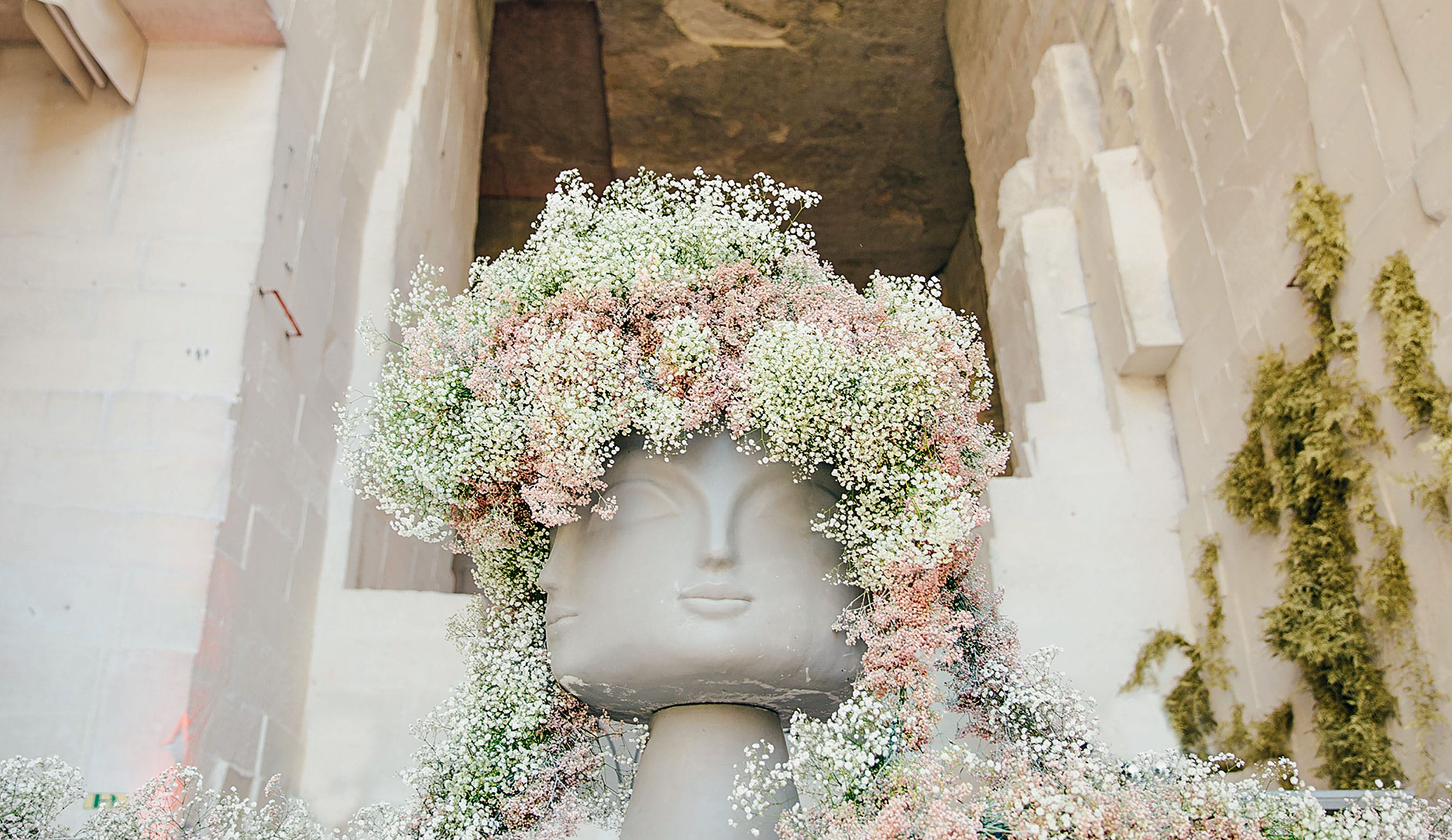 Sculpted head adorned with cascading pink and white flowers, set against a textured stone background.