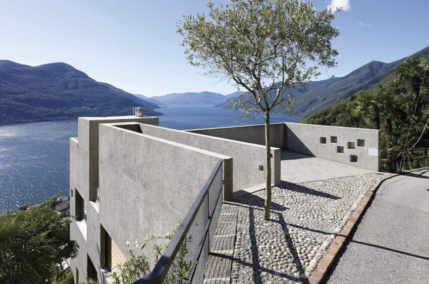 Modern concrete structure with a tree on a terrace overlooking a scenic lake and mountainous landscape under a clear blue sky.