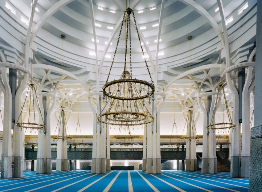 Interior of a mosque with large chandeliers, white pillars, and blue carpet rows forming a striking symmetrical design.