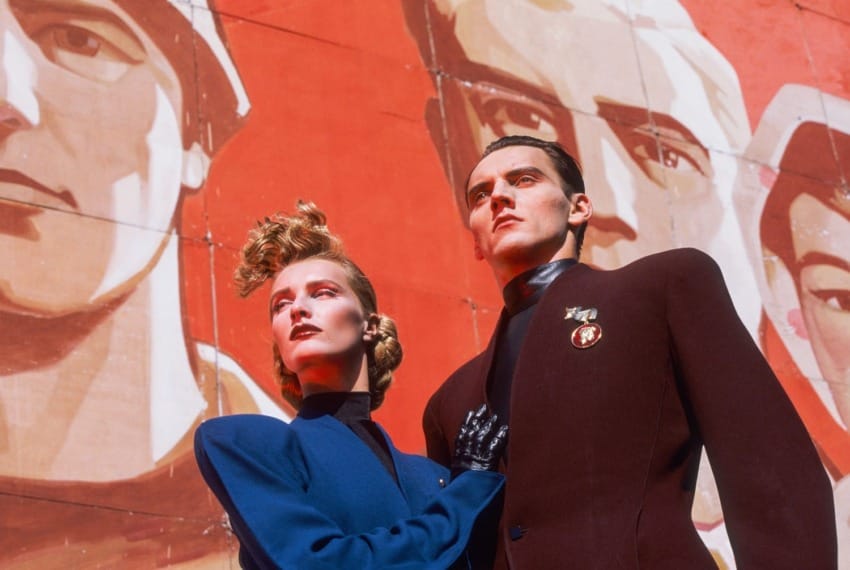 Stylishly dressed couple in front of a large, red mural featuring oversized faces in a bold, dramatic setting.