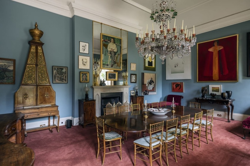 Elegant dining room with ornate chandelier, vintage furniture, and various artworks on blue walls.