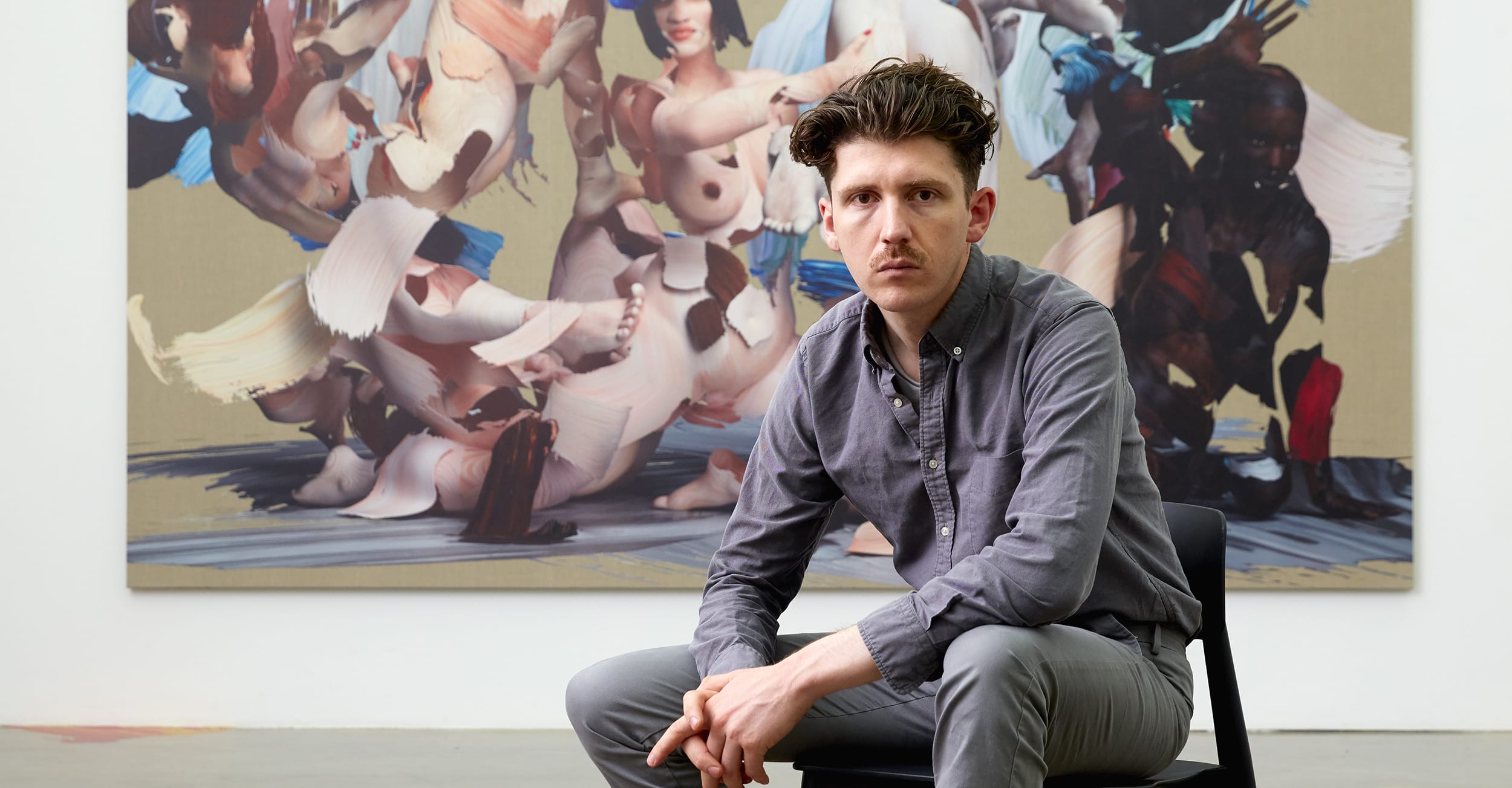 Man seated in front of a large abstract painting, wearing a gray shirt and pants, in a modern art gallery setting.