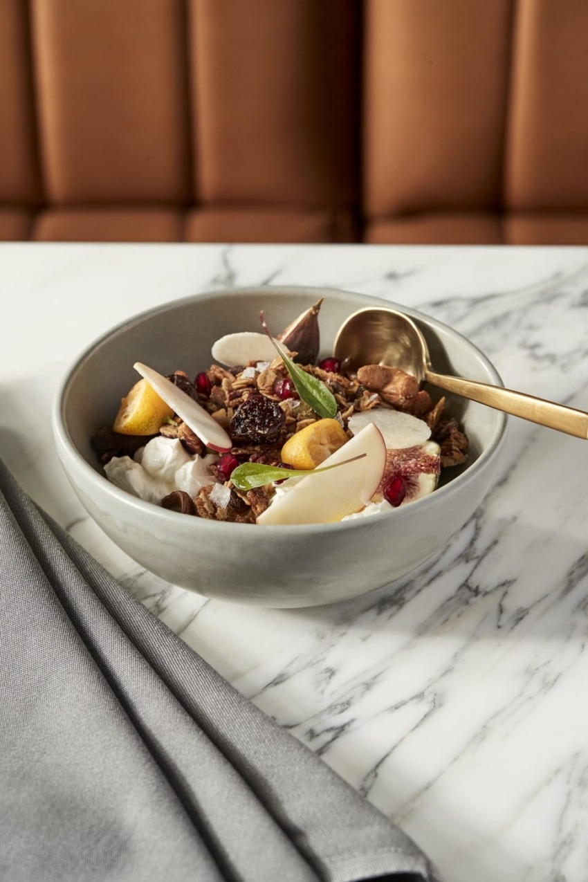 Bowl of yogurt with granola, pear slices, nuts, and a spoon on a marble table with gray napkin and cushioned seating background.
