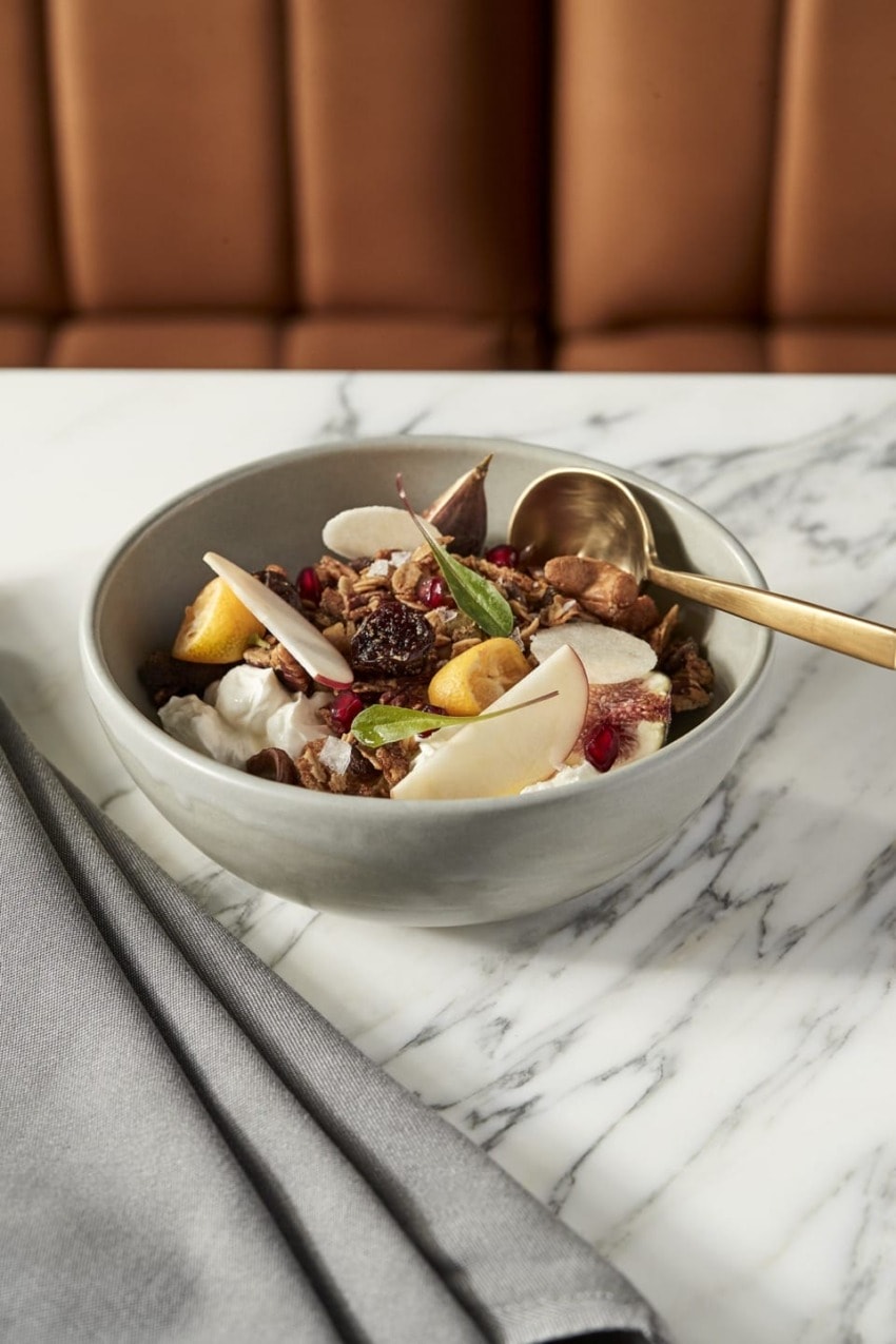 Bowl of yogurt with granola, pear slices, nuts, and a spoon on a marble table with gray napkin and cushioned seating background.