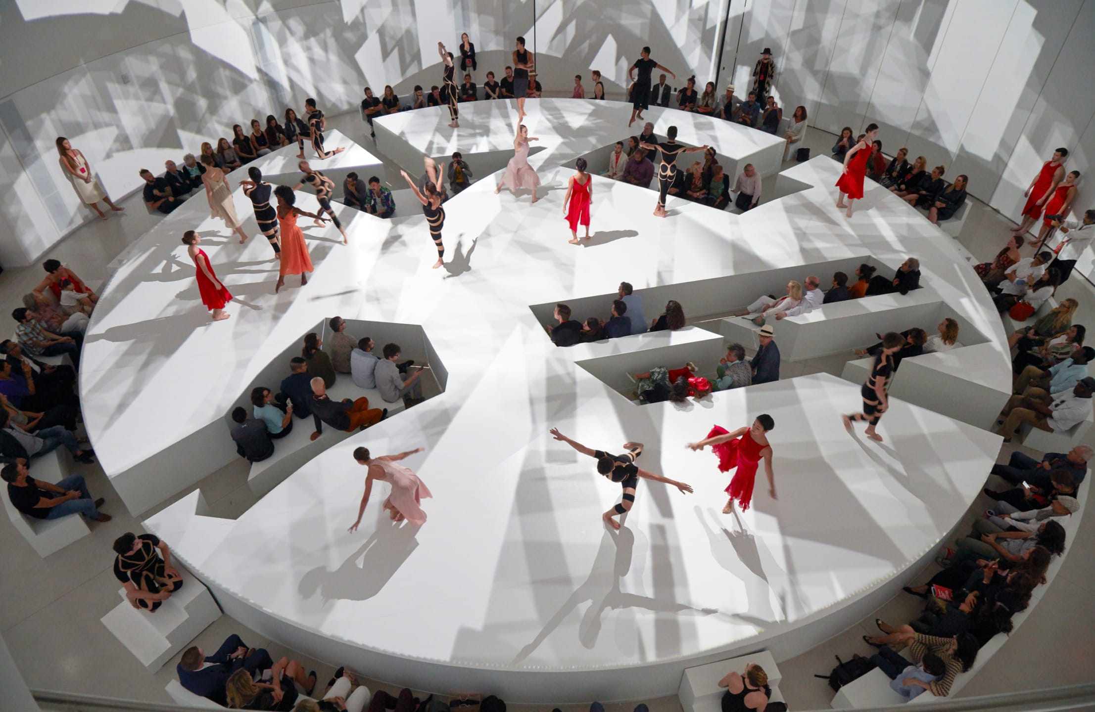Models in colorful dresses on a circular runway with audience seated around, casting artistic shadows on white surface.