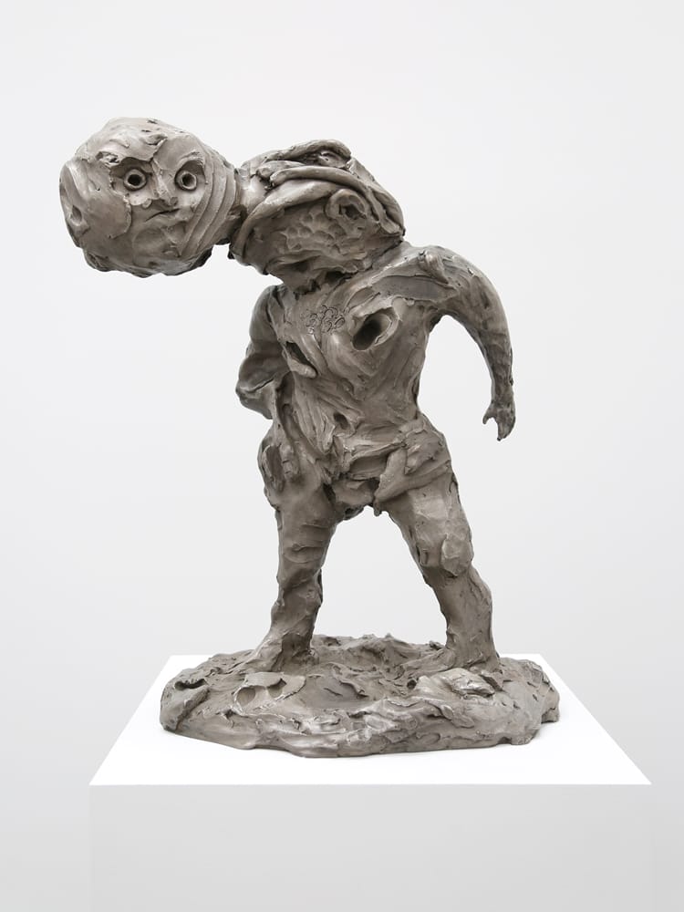 Contemporary abstract sculpture of a humanoid figure with a distorted head, displayed on a white pedestal in a gallery setting.