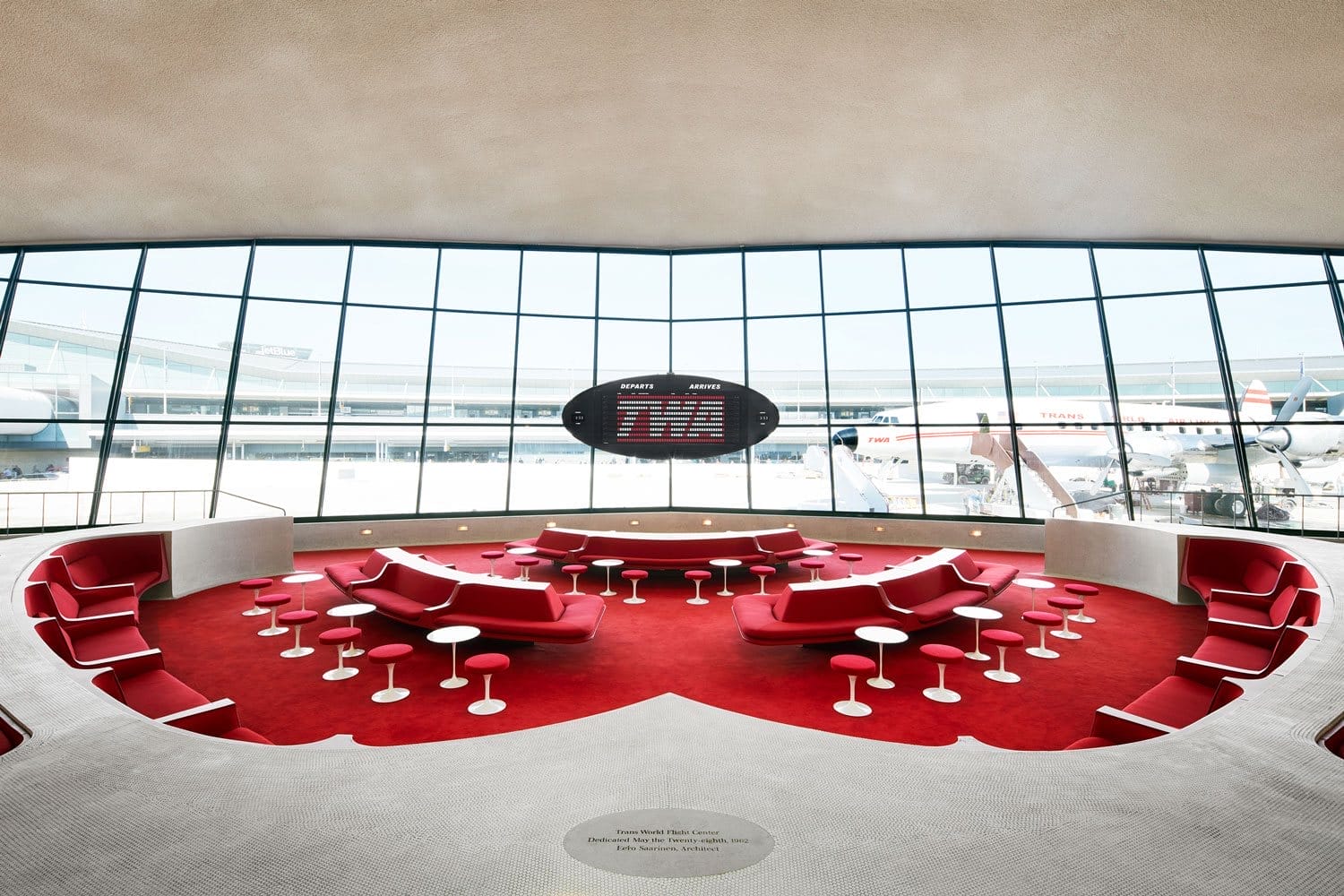 Retro-style airport lounge with red seating, round tables, and a panoramic window overlooking the runway and parked airplanes.