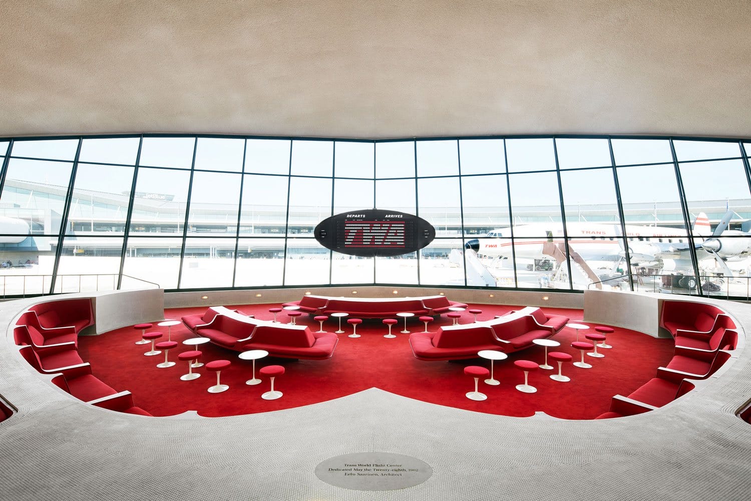 Retro-style airport lounge with red seating, round tables, and a panoramic window overlooking the runway and parked airplanes.