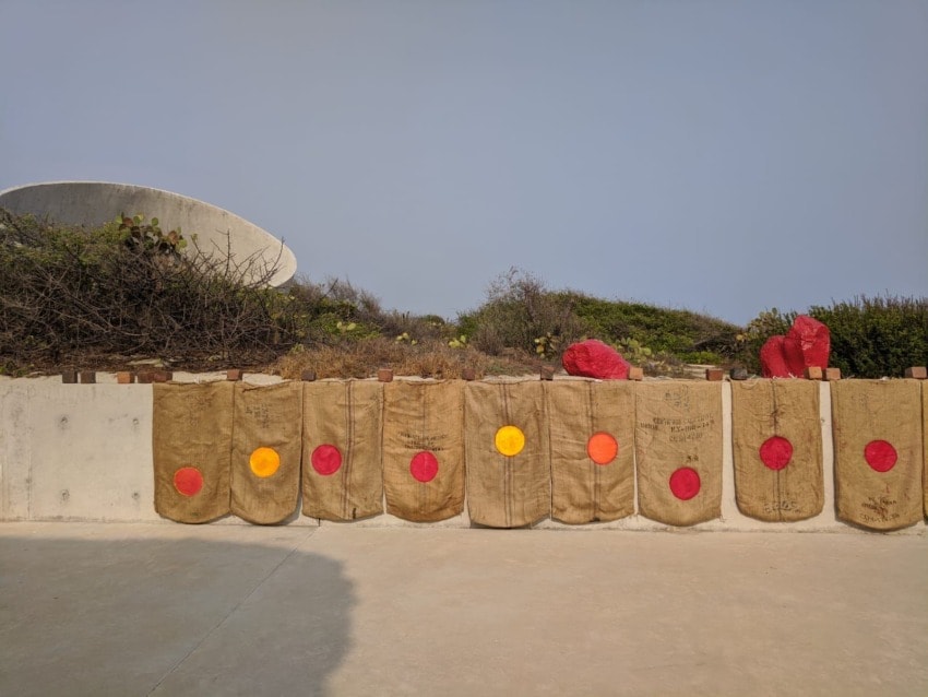 Outdoor art display with burlap bags adorned with colorful circles against a concrete wall and shrubs.