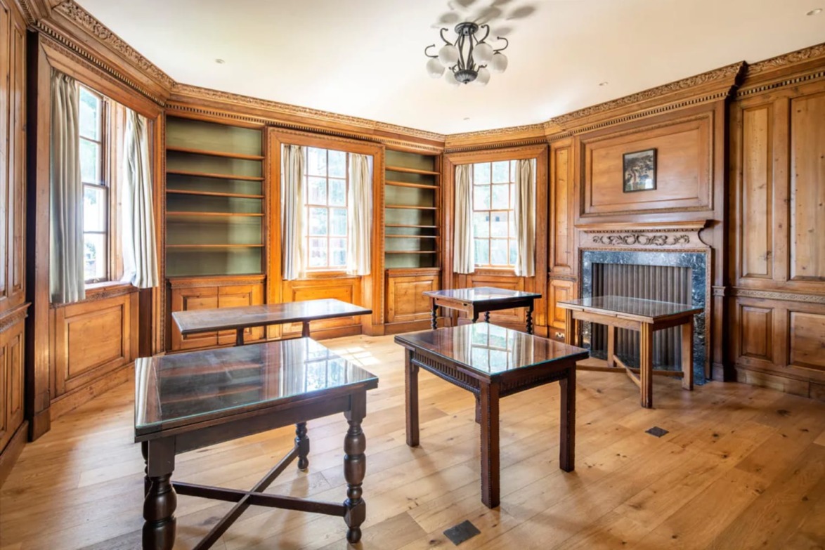 Elegant library room with wooden paneling, several glass-topped tables, large windows, and an ornate fireplace.