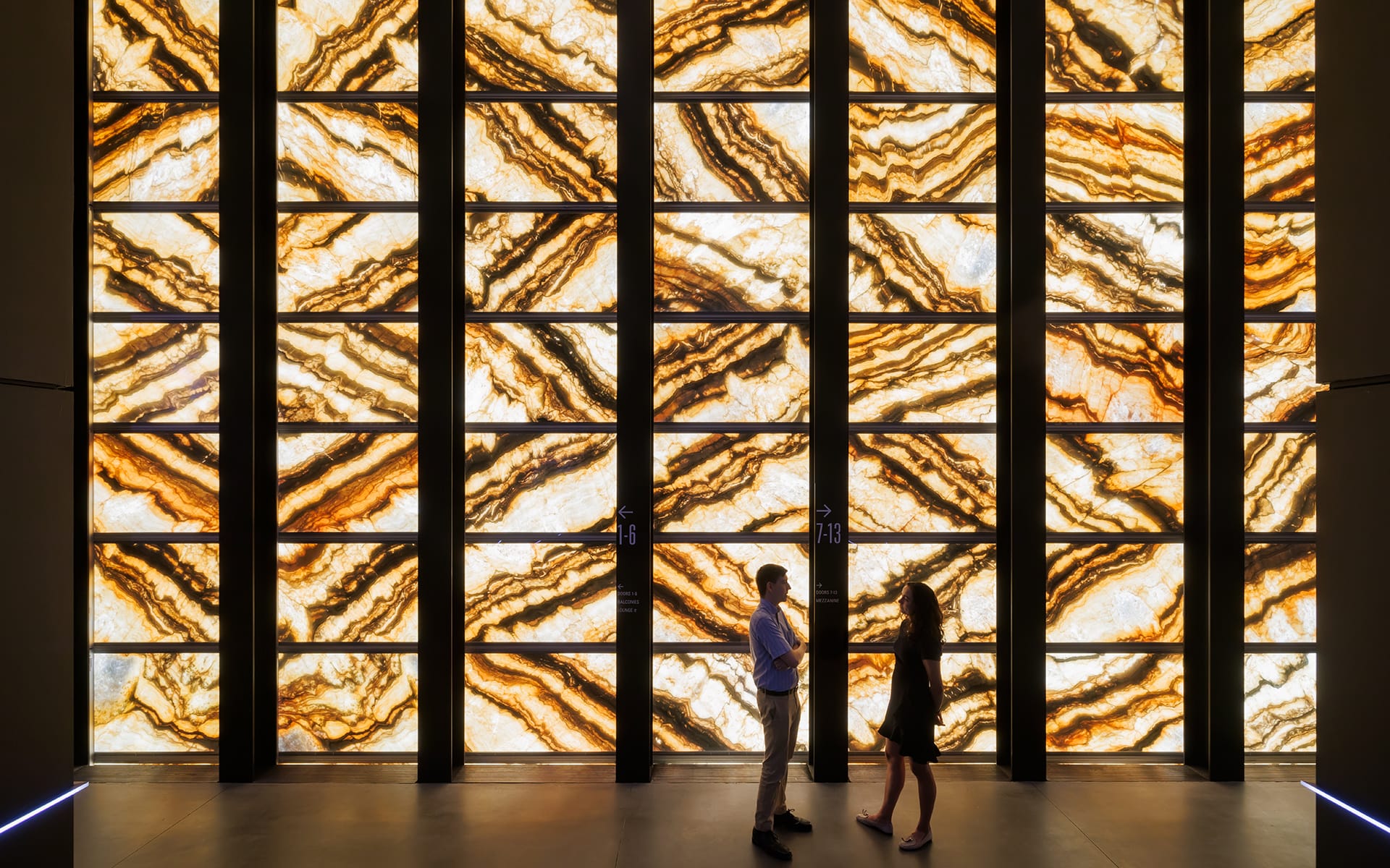 Silhouettes of two people talking in front of a large backlit onyx wall with intricate patterns.