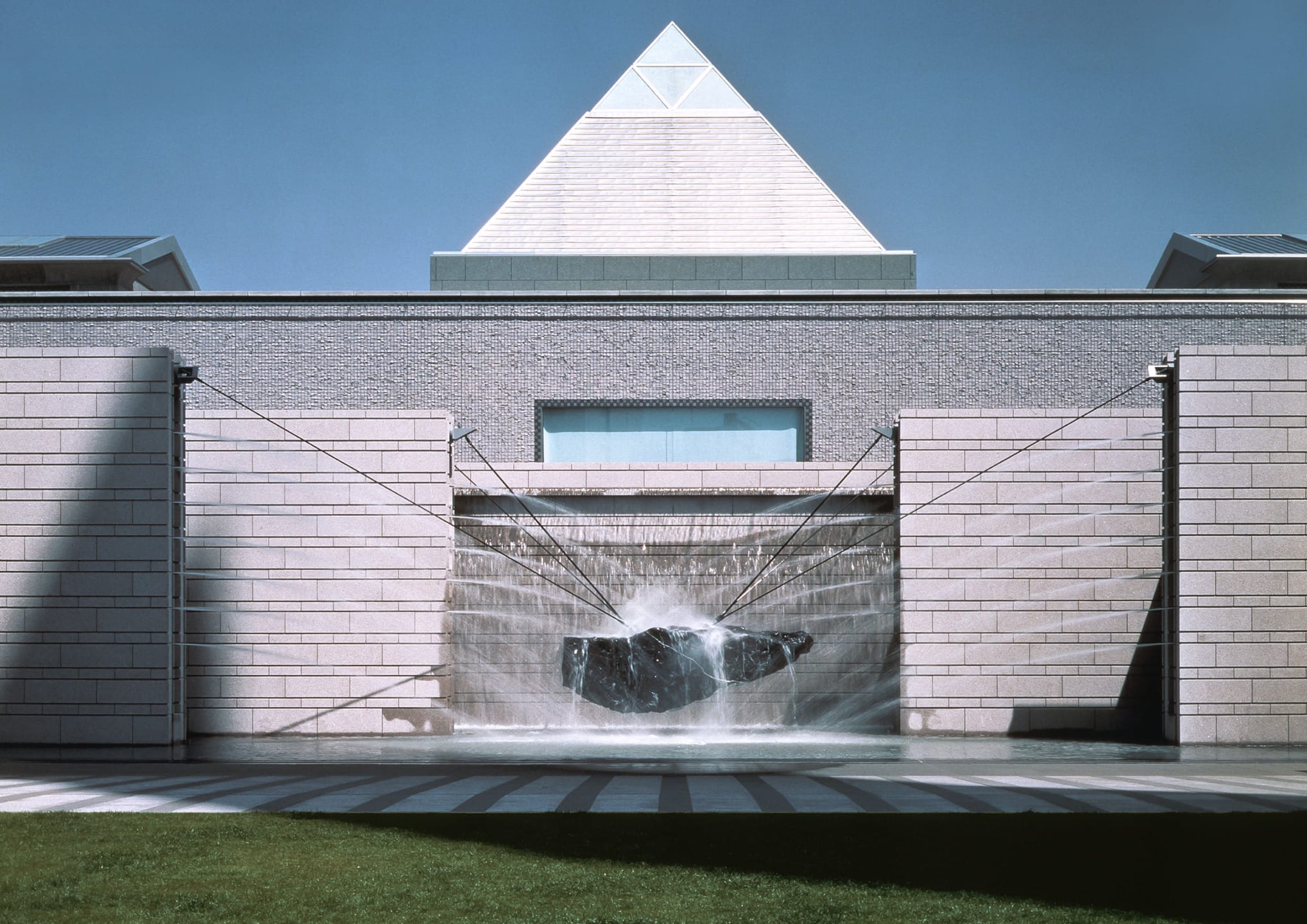 Fountain with suspended rock in front of a modern building with a pyramid-shaped glass roof, against a clear blue sky.