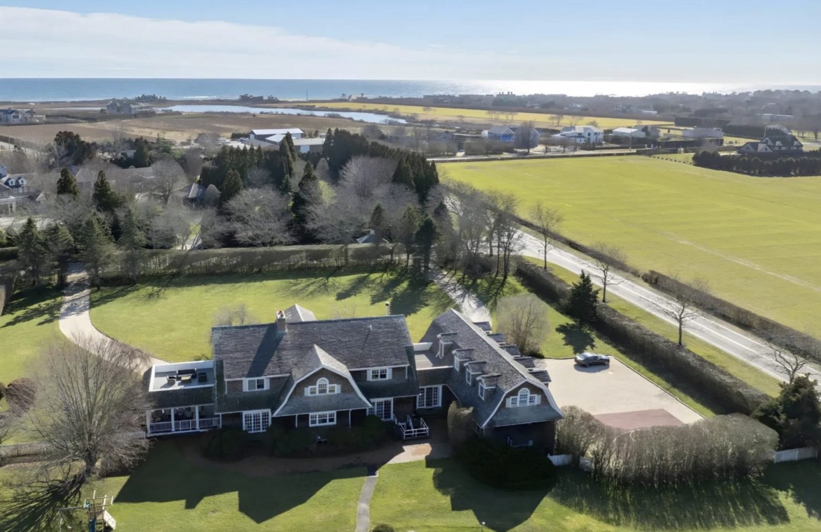 A large house with a gray roof surrounded by fields and trees, near the ocean and patches of water visible in the distance.