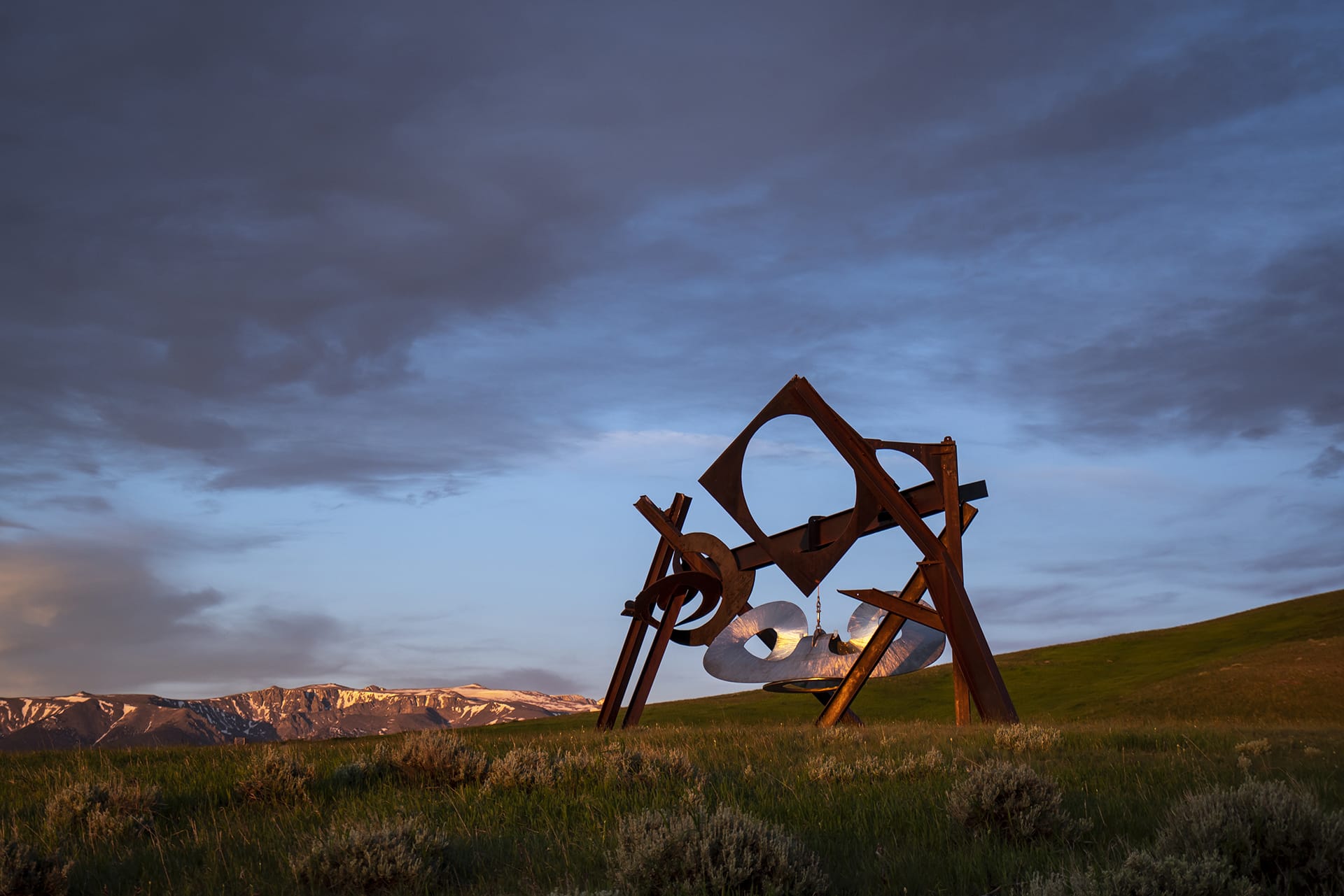 Abstract metal sculpture on grassy landscape with mountains and dramatic cloudy sky in the background.