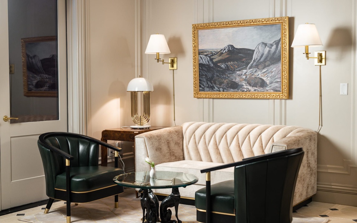 Elegant living room with velvet sofa, two green chairs, glass table, wall painting, and stylish lighting fixtures.