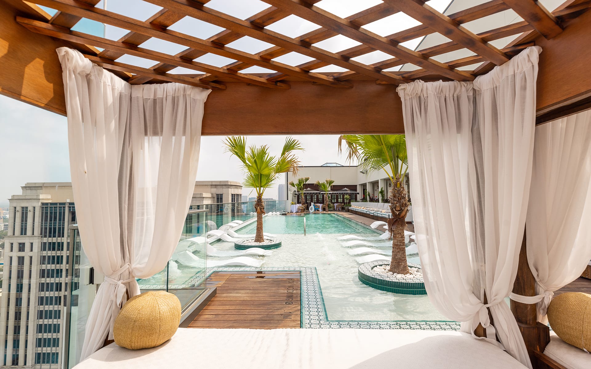 Rooftop pool with lounge chairs and palm trees, framed by white curtains under a wooden pergola on a sunny day.