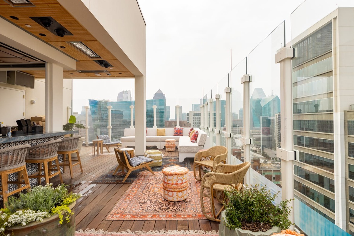 Rooftop patio with city view, featuring lounge seating, decorative rugs, and bar area with wooden stools and plants.