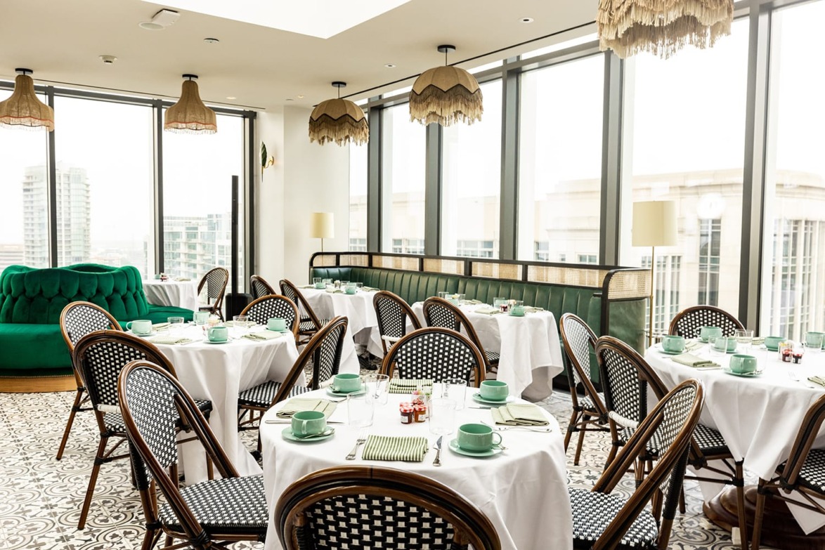 Stylish dining area with wicker chairs, white tablecloths, and large windows offering a city view.