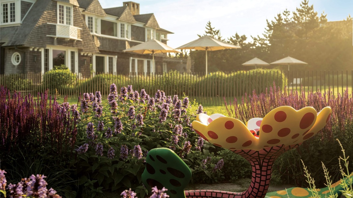 Garden with vibrant flowers and an artistic sculpture in front of a large house with a sunlit backdrop.