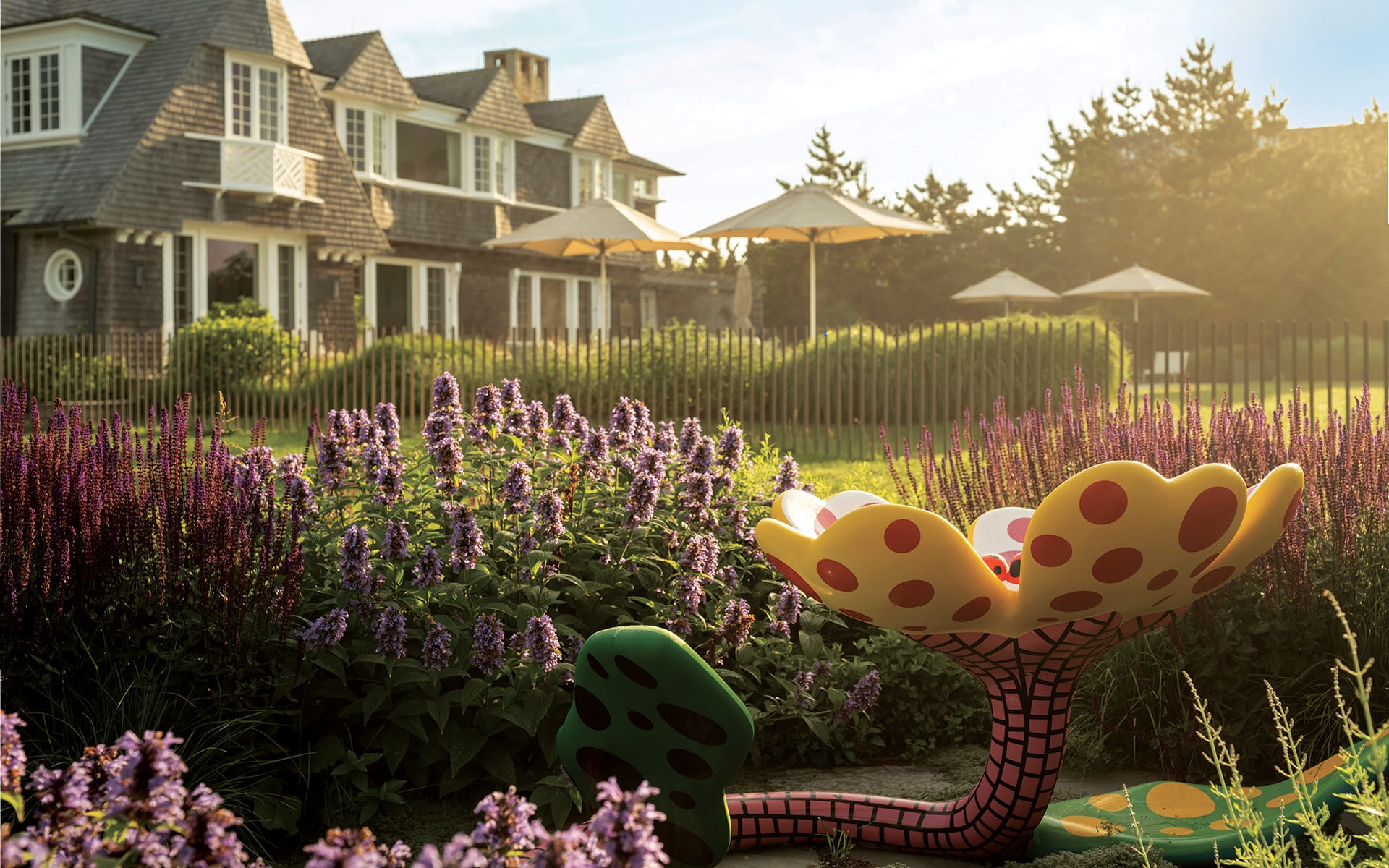 Garden with vibrant flowers and an artistic sculpture in front of a large house with a sunlit backdrop.