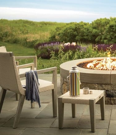 Outdoor seating by a fire pit with a thermos on a table, surrounded by greenery and a meadow in the background.