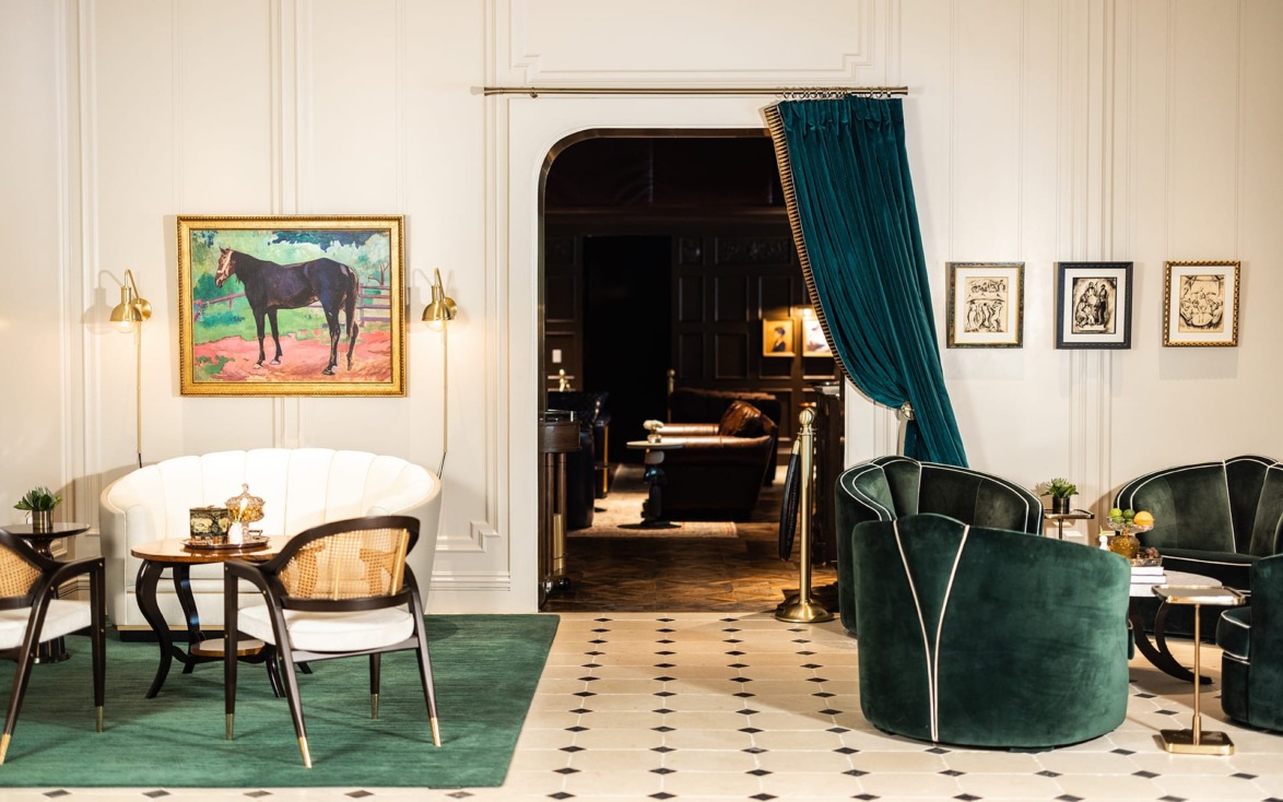 Elegant lounge with green velvet chairs, artwork on white walls, and a partly open curtain revealing a cozy room beyond.