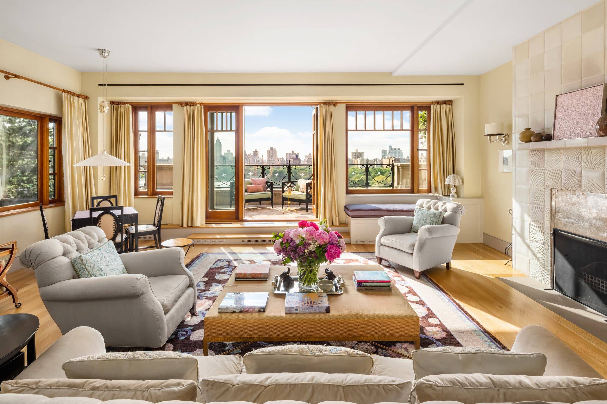 Spacious living room with large windows, elegant furniture, a coffee table with flowers, and a view of a cityscape.