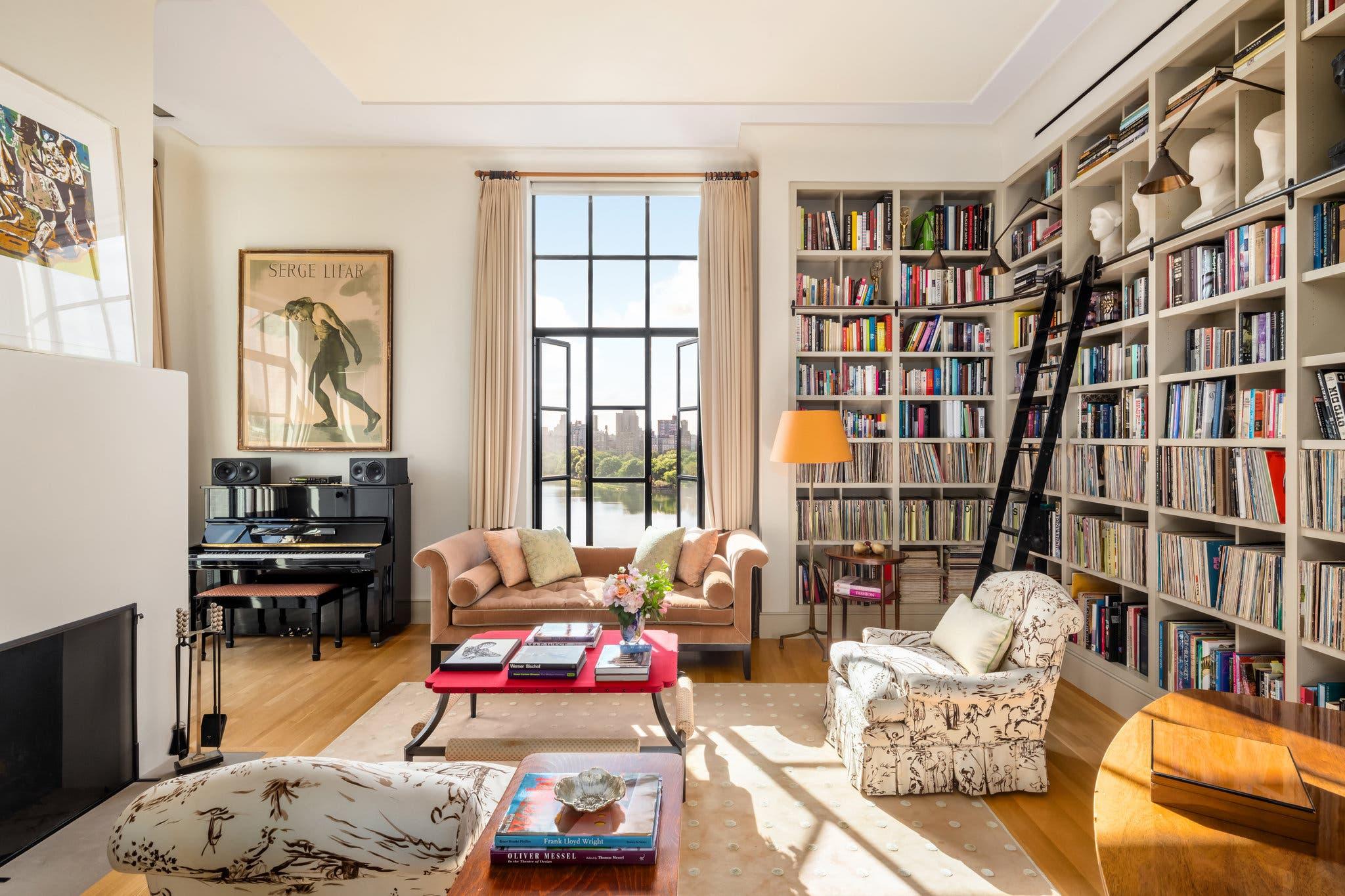 Cozy living room with bookshelves, a grand piano, sofa, armchairs, large windows, and sunlight streaming in.
