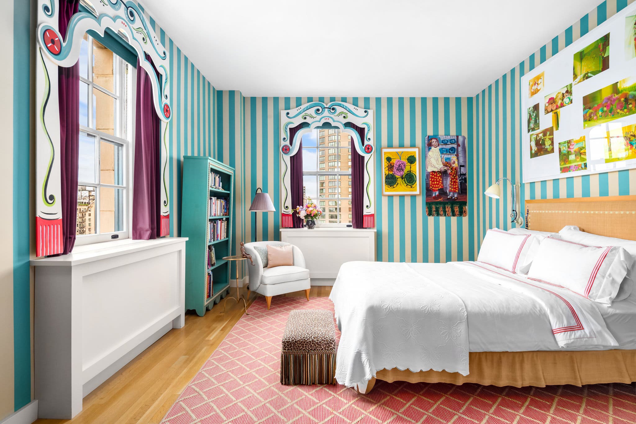 Bright bedroom with striped teal walls, colorful art, double windows, white bedding, and a cozy armchair by the radiator.