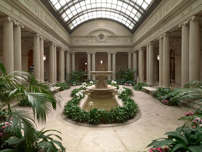 Elegant indoor garden with central fountain surrounded by green plants and flowers, under a large glass dome ceiling.