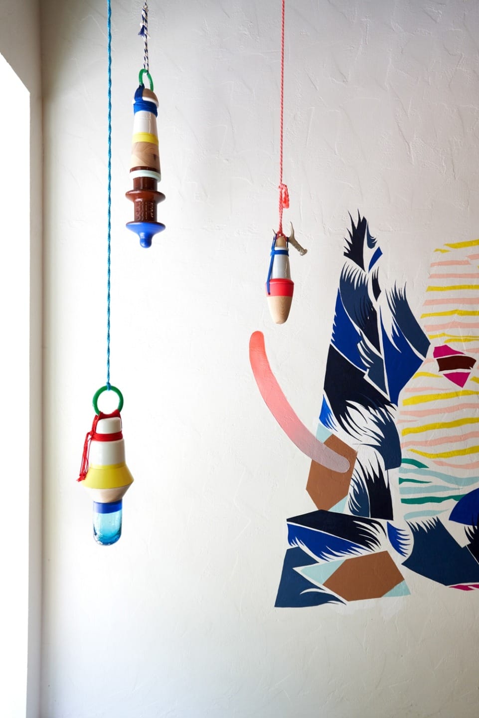 Colorful hanging ornaments and a vibrant abstract wall mural on a textured white wall, with natural light from a window.