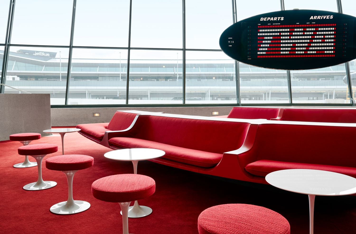 Retro airport lounge with red seating, round tables, and a vintage arrivals and departures board in the background.