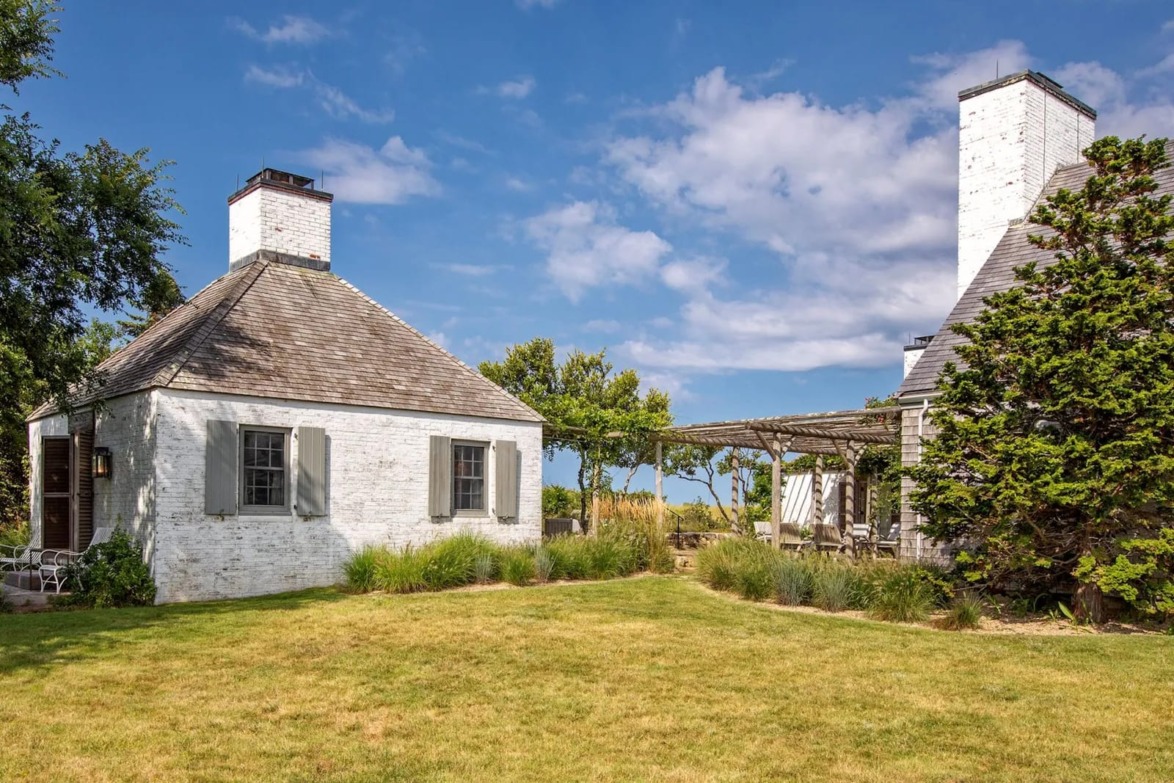 Two white brick cottages with lush green grass, trees, and a sunny blue sky in the background.