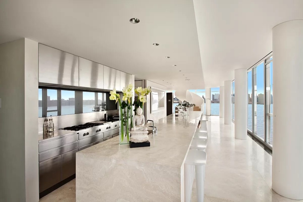 Modern kitchen with marble countertops, sleek appliances, white stools, and large windows overlooking a waterfront view.