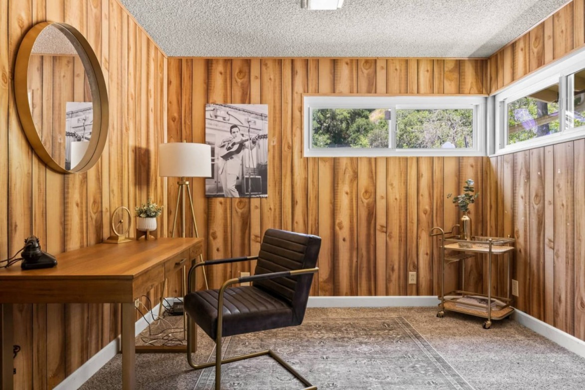 wood-paneled office with a wooden desk, black chair, round mirror, and a window with a view of greenery