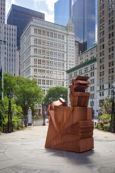 Rust-colored geometric sculpture in urban park setting, surrounded by tall buildings and greenery.