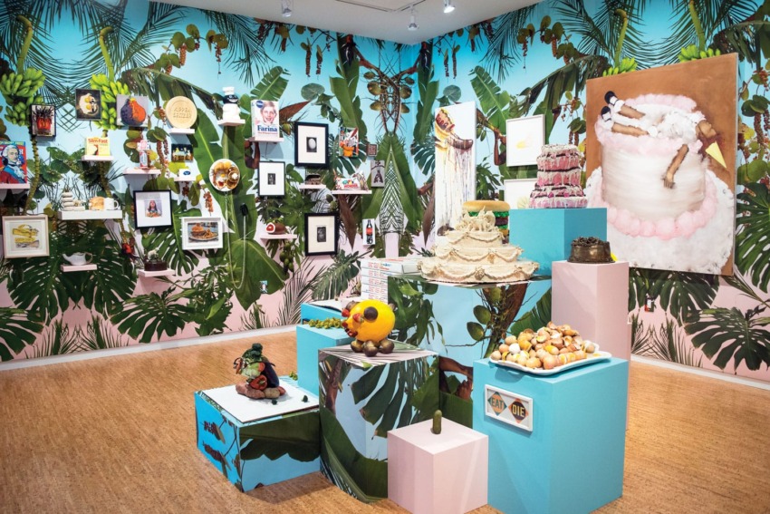Tropical-themed art exhibit with various paintings and sculptures on pedestals in a vibrant, colorful gallery space.