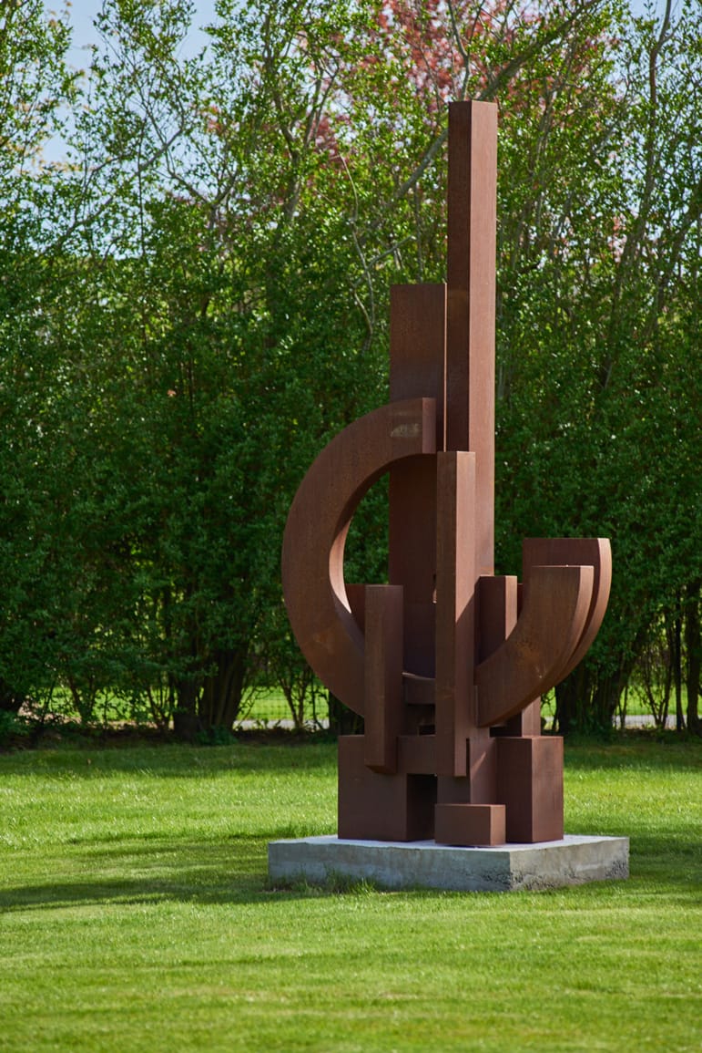 modern rust-colored abstract sculpture on green lawn with trees in background