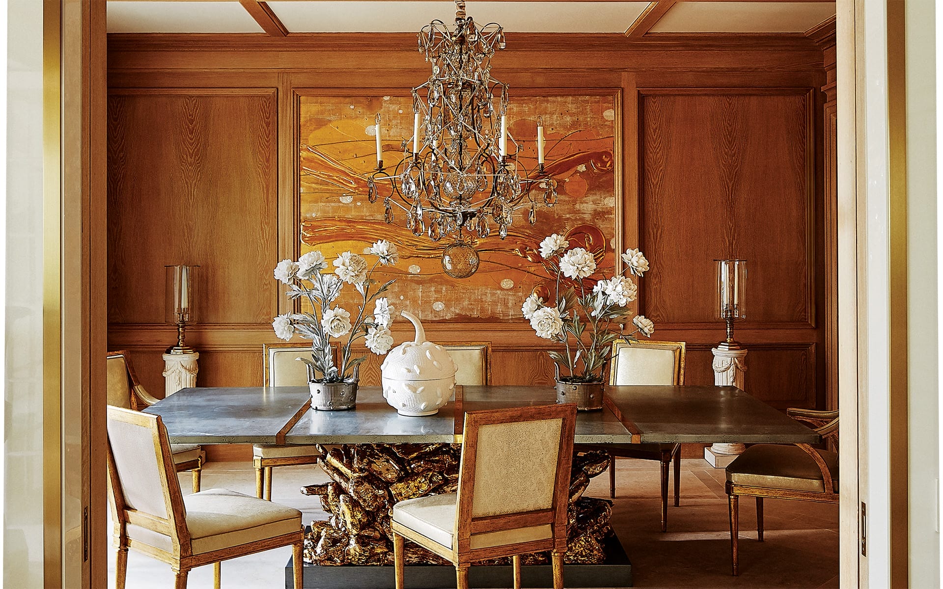 Elegant dining room with chandelier, modern art, and wooden table surrounded by upholstered chairs and decorative vases.