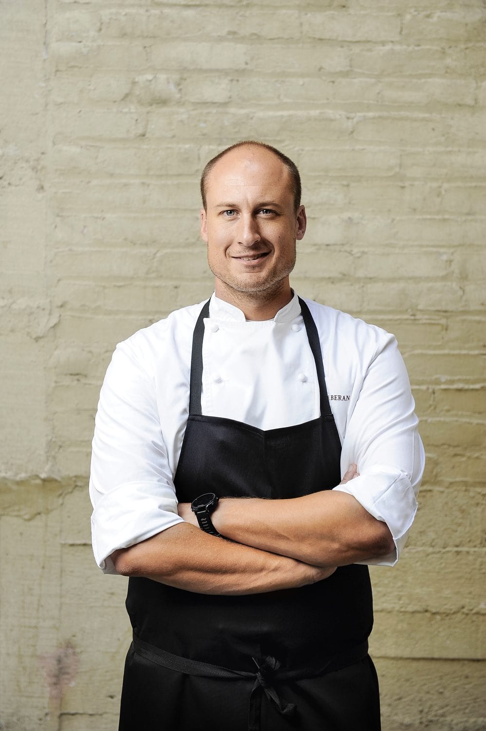 Chef in a black apron over a white chef's jacket standing with arms crossed against a textured beige wall.