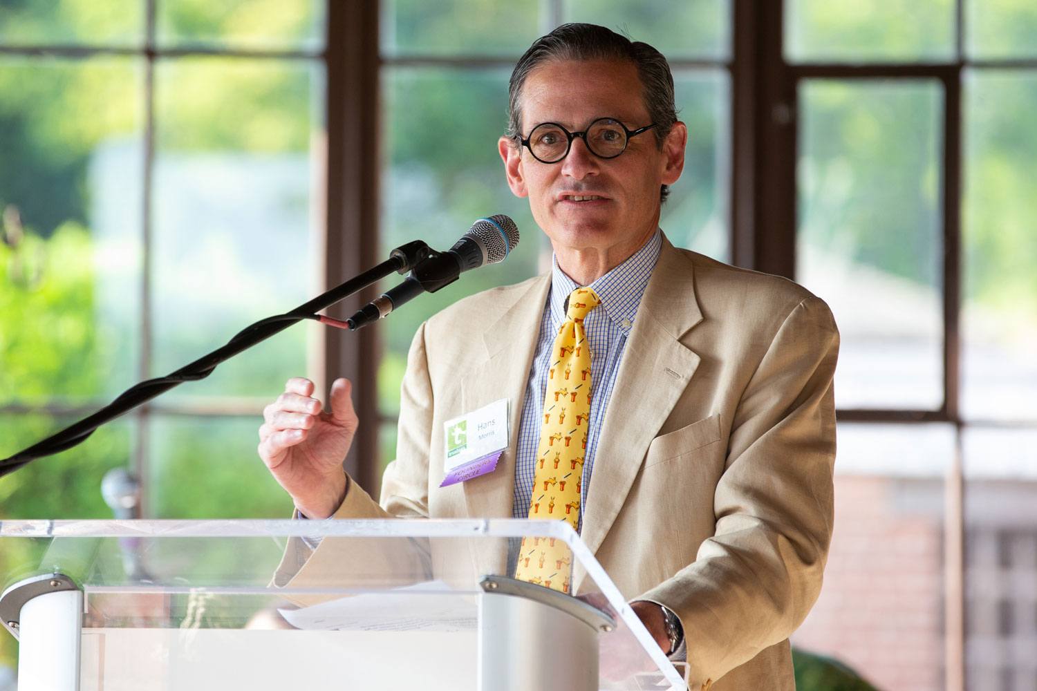 Man with glasses and a tan suit speaking at a podium with a microphone in front of a windowed background.
