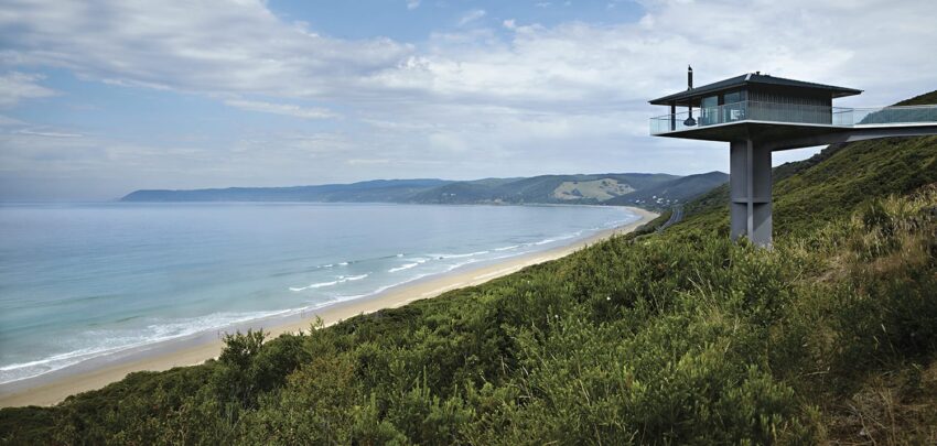 Modern house on elevated platform overlooking a scenic coastline with lush greenery and a clear blue sky above the ocean.