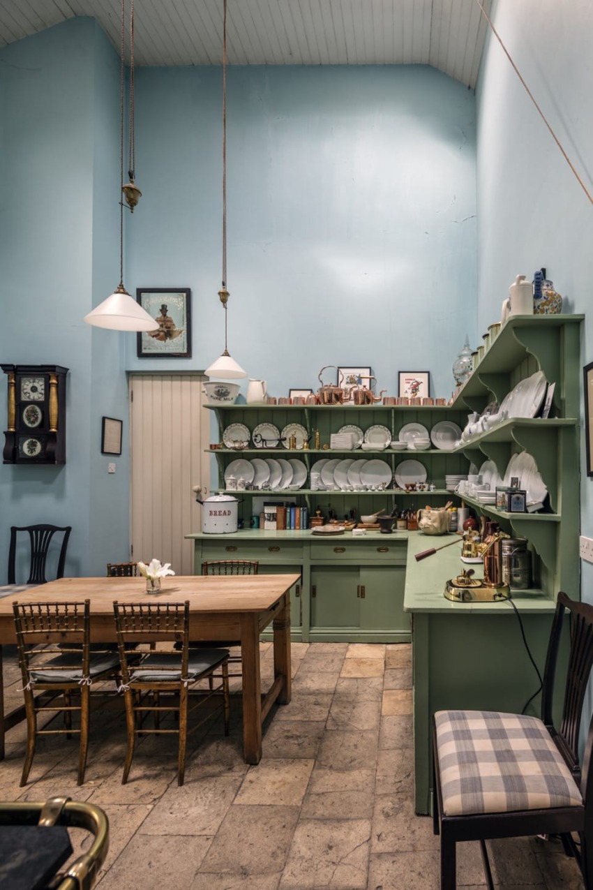 Vintage kitchen with light blue walls, wooden table, hanging lights, green shelves, and vintage decor.