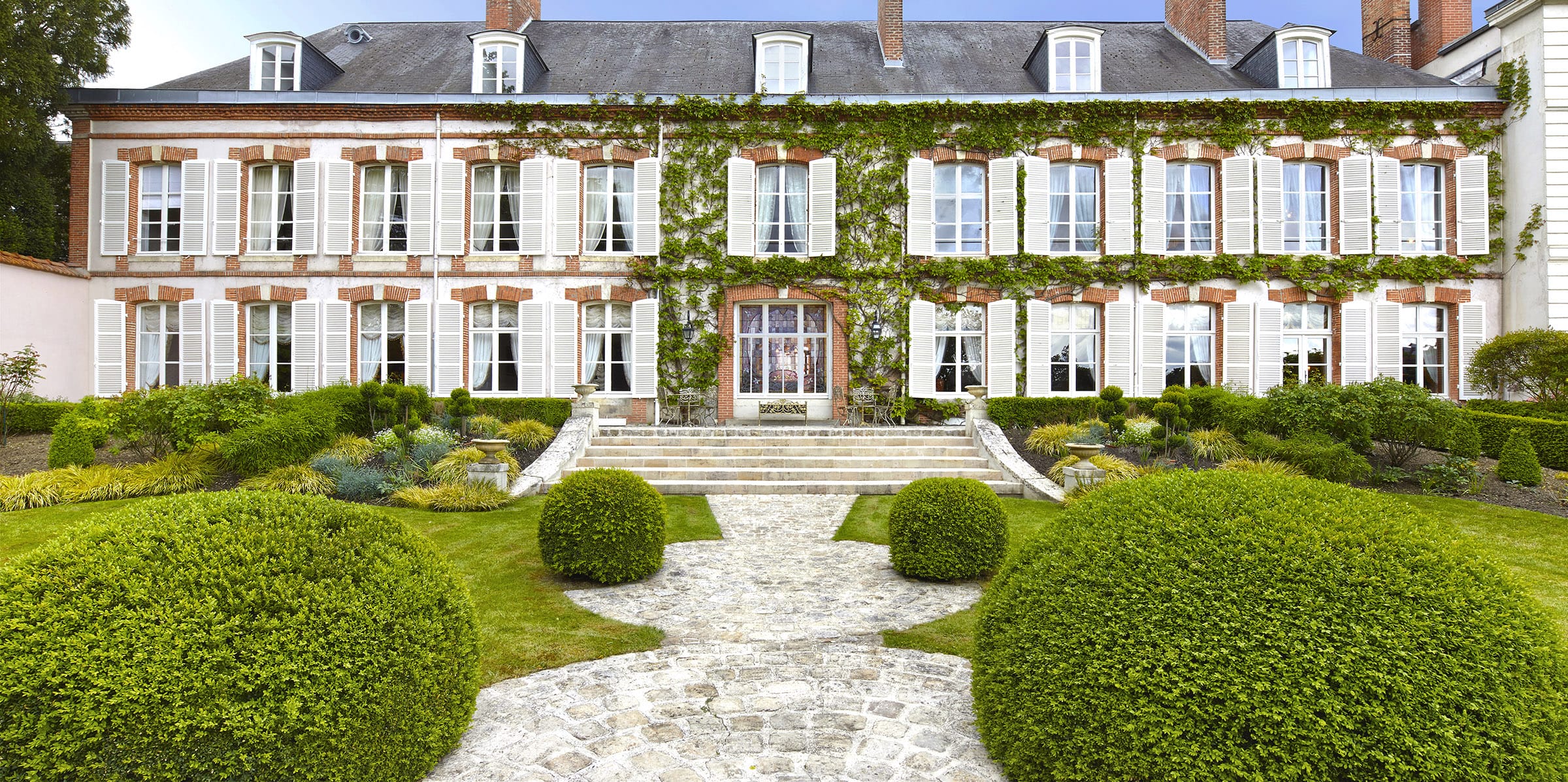 Elegant historic mansion with ivy-covered walls, centered doorway, symmetrical windows, and a formal garden featuring round shrubs.